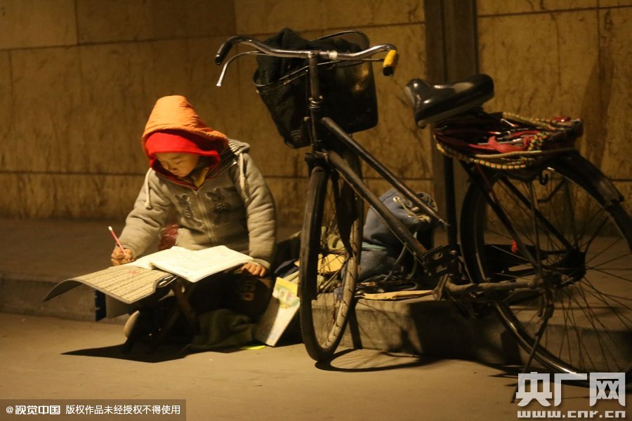 鄭州中原路京廣鐵路大橋下涵洞的馬路牙子邊，一位戴著紅色辣椒帽的小男孩趴在一個(gè)小馬扎上，上面鋪著一張車墊，在微弱的燈光下寫作業(yè)